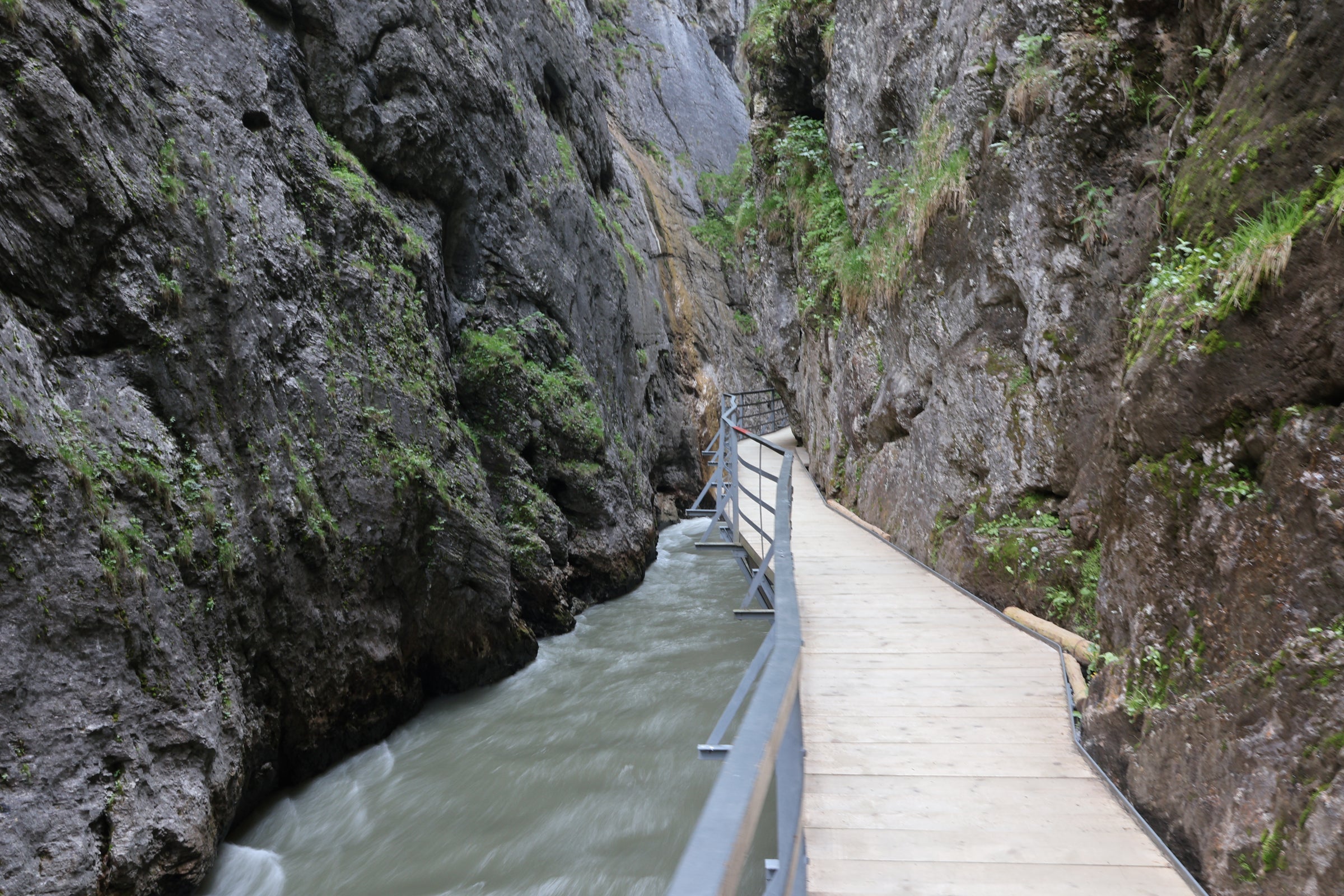 A wooden boardwalk with metal railings runs alongside a narrow river flowing between steep rocky canyon walls with patches of green moss and vegetation.