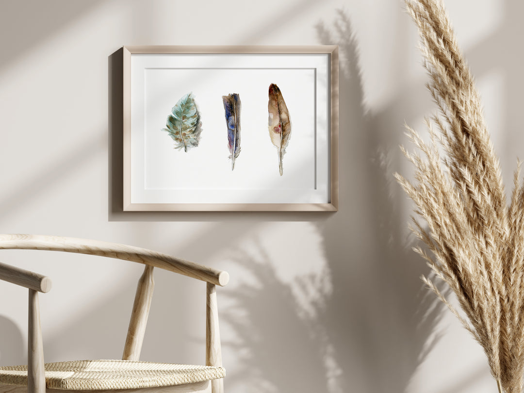 Framed artwork of feathers on a wall with a chair and dried plants in the foreground