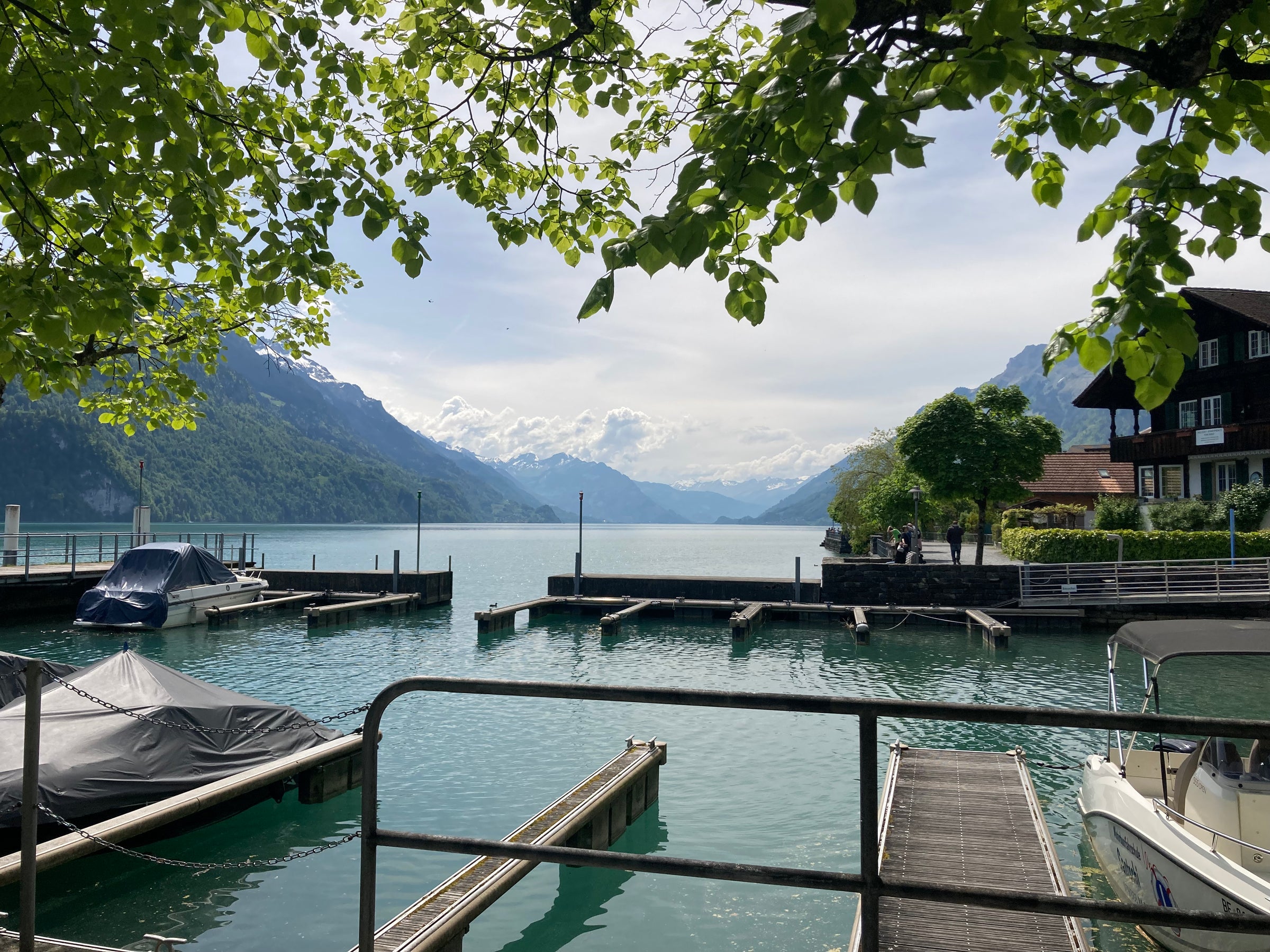 A scenic view of a calm Swiss lake with wooden docks, covered boats, a traditional alpine-style house on the right, and mountains in the background under a cloudy sky, framed by green tree leaves in the foreground.