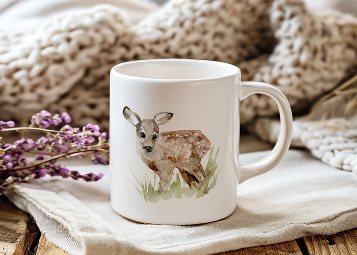 White mug with a deer illustration on a wooden surface with a textured blanket and purple flowers in the background.
