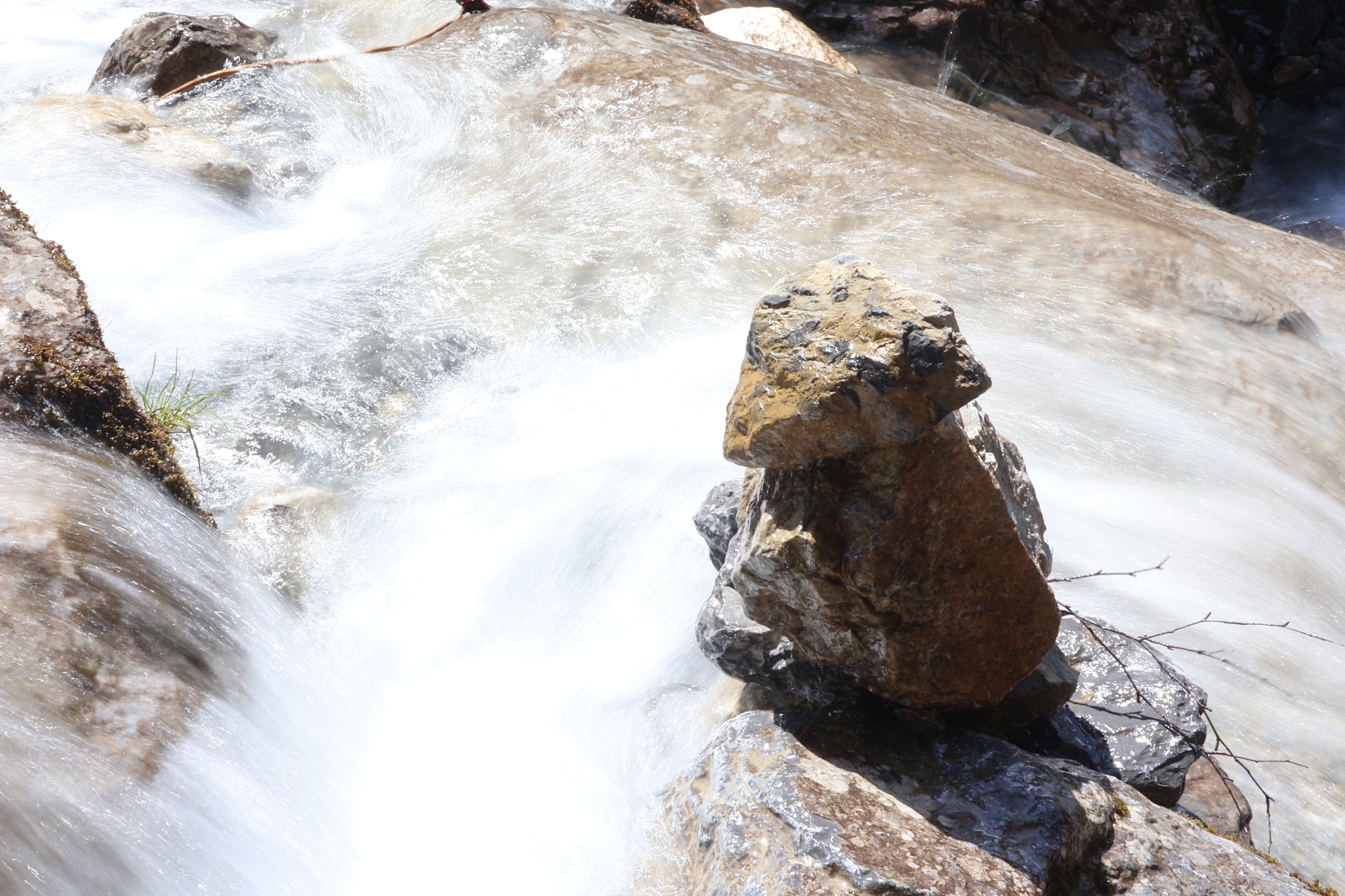 A close-up outdoor scene of rushing water over stacked rocks, with a prominent brown and gray boulder in the foreground and white water filling much of the frame.