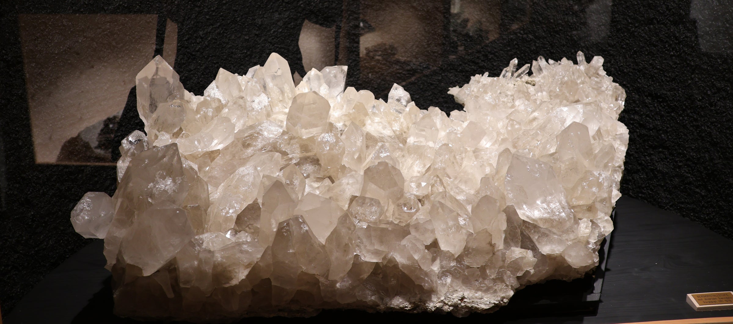 A large natural quartz crystal cluster specimen with clear, faceted white quartz points, displayed on a dark surface against a dark background.