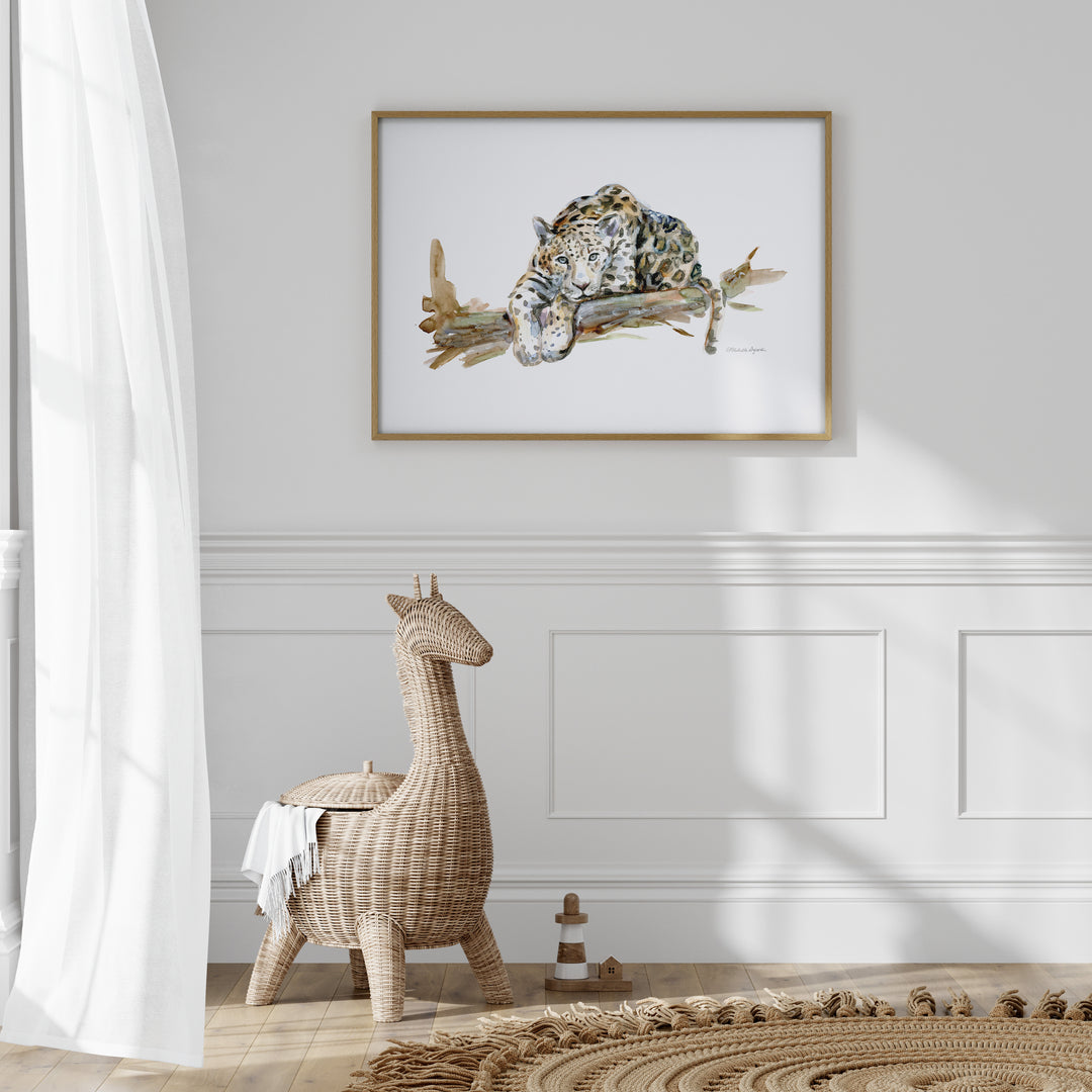 Framed artwork of a jaguar on a branch above a wicker stool in a room with white walls and wooden flooring.