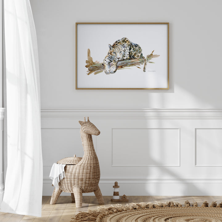 Framed artwork of a jaguar on a branch above a wicker stool in a room with white walls and wooden flooring.