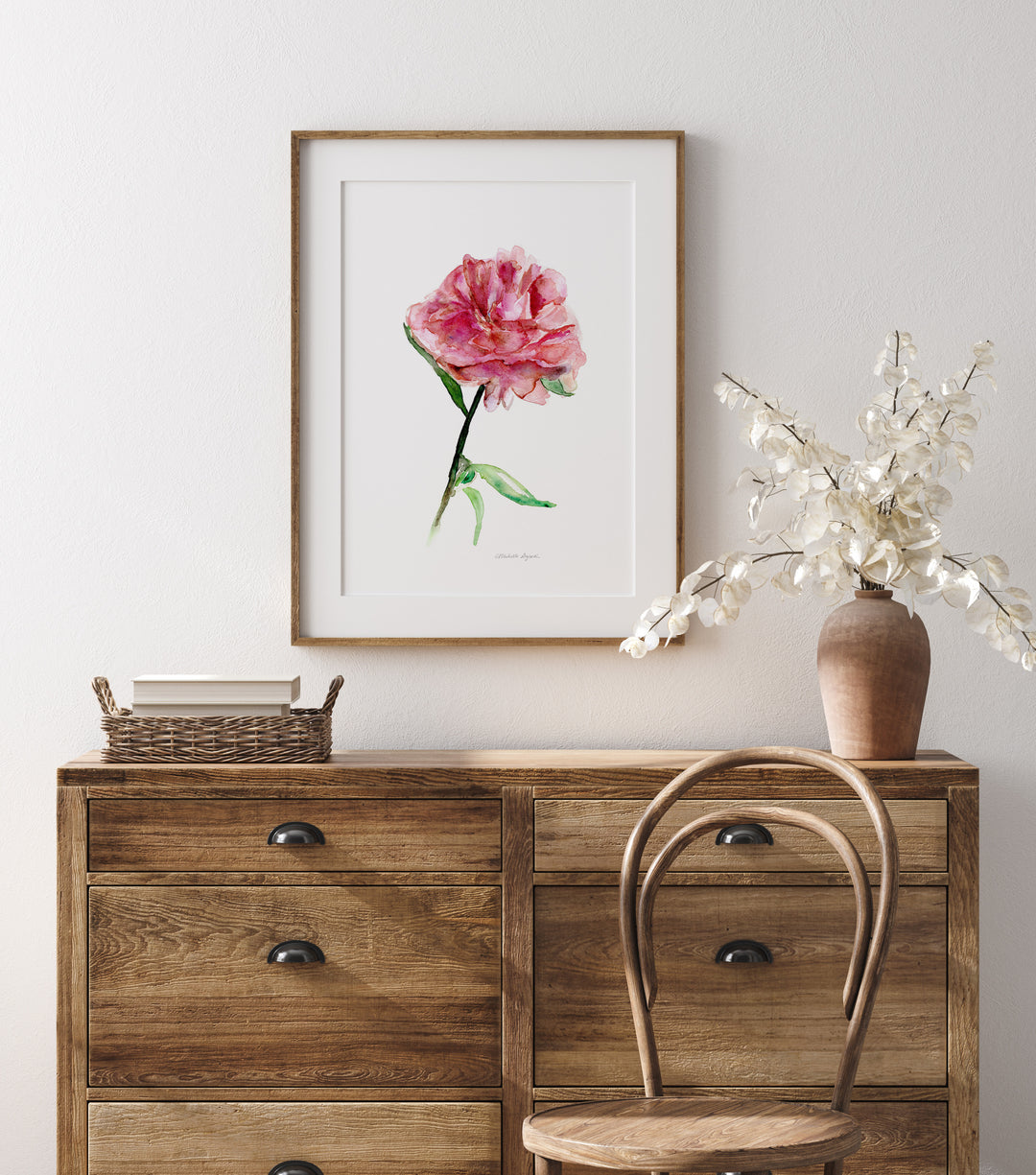 Wooden dresser with a framed pink flower print and a vase with white flowers on a light gray wall.